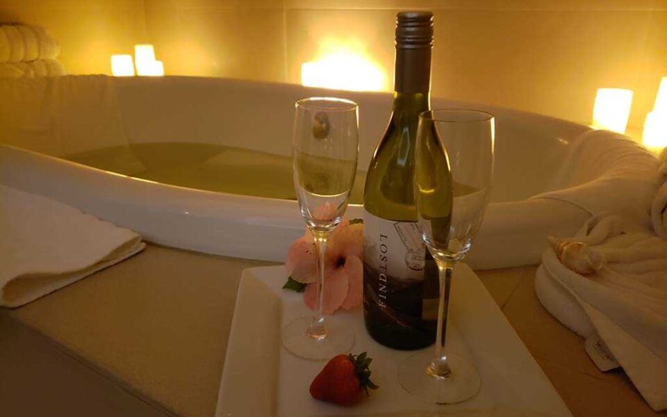 a bottle of wine and two glasses sit by a full bathtub surrounded by glowing candles